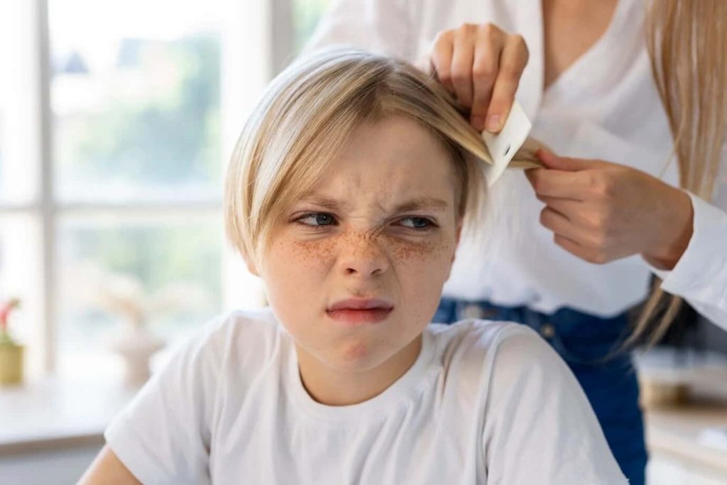 Un enfant blond grimace pendant une séance de peignage avec un peigne fin, évoquant un traitement efficace dans un centre anti poux.