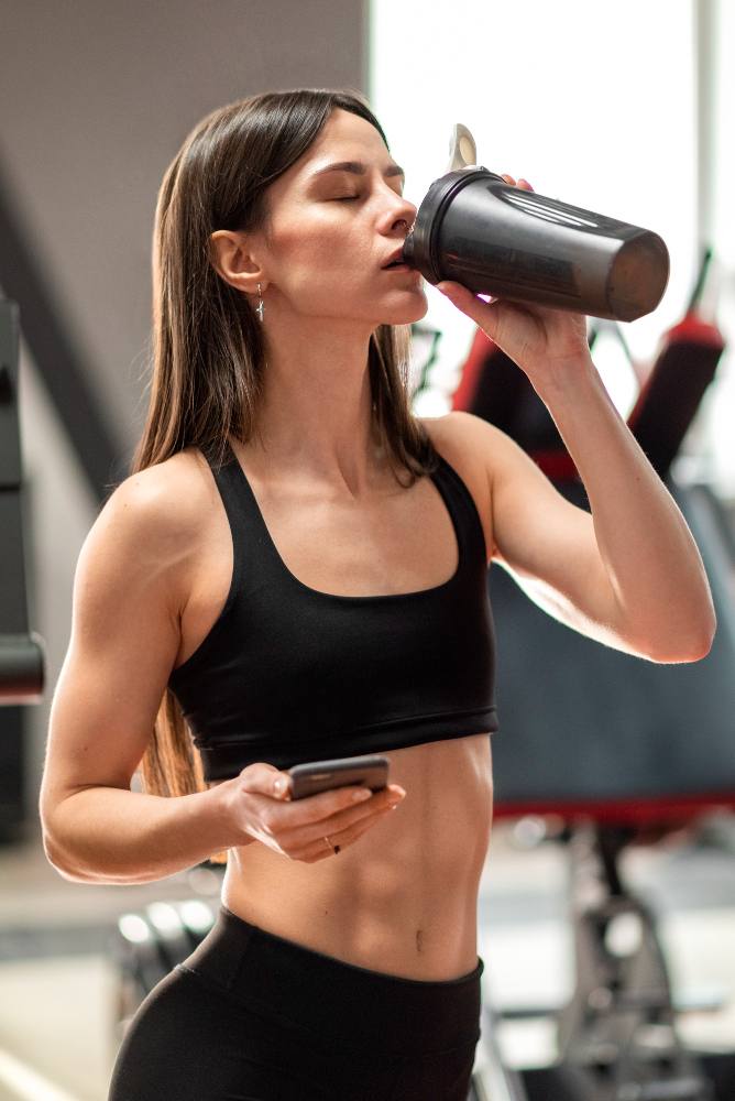 Une femme athlétique en brassière et legging noirs boit dans un shaker en plastique tout en tenant son smartphone dans une salle de sport.
