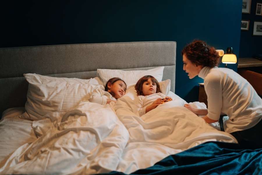 Une mère bordant ses deux enfants dans un grand lit douillet, un moment clé pour instaurer une routine du coucher apaisante.